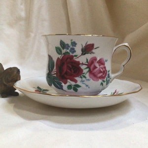 May include: A white teacup and saucer with a floral pattern featuring pink and red roses, blue flowers, and green leaves. The cup and saucer have a gold trim.