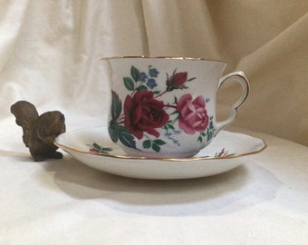 May include: A white teacup and saucer with a floral pattern featuring pink and red roses, blue flowers, and green leaves. The cup and saucer have a gold trim.