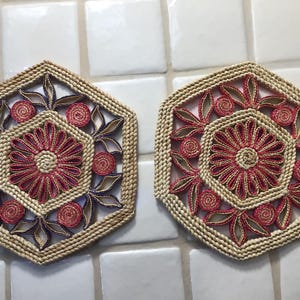 Puede incluir: Dos salvamanteles hexagonales tejidos. Cada salvamanteles presenta un diseño floral central en rojo y beige, rodeado de hojas estilizadas y acentos en espiral. Los bordes son de color beige natural, y los salvamanteles están colocados sobre una superficie de baldosas blancas.