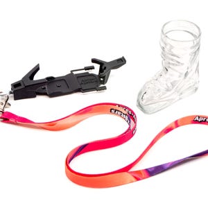 May include: A black ski boot binding, a clear glass boot-shaped shot glass, and a lanyard with a red and orange gradient design. The lanyard has the text "Apres-Allstars" printed on it. The items are on a white background.