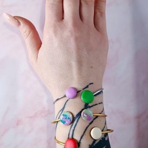 Può includere: Quattro braccialetti con accenti rotondi e colorati sono esposti su un polso. I braccialetti hanno fasce dorate e argentate. Gli accenti sono nelle tonalità di rosa, verde, rosso e bianco, alcuni con un design marmorizzato.