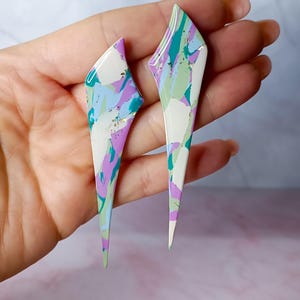 Può includere: Un paio di orecchini astratti e geometrici. Ogni orecchino ha una forma lunga e sottile con un'estremità a punta. Gli orecchini presentano un motivo marmorizzato di bianco, verde acqua, lavanda e accenti dorati. Gli orecchini sono tenuti in una mano.