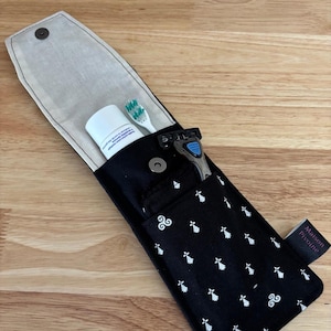 Peut inclure: Trousse de toilette de voyage noire avec motif floral blanc, contenant une brosse à dents, un rasoir et un déodorant. Le sac a une fermeture à bouton-pression et un intérieur clair. L'étiquette indique "Maison Pivoine".