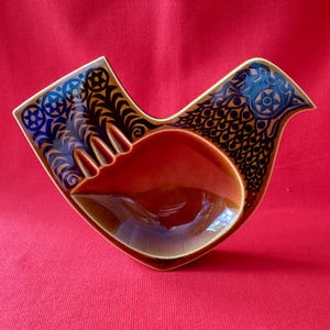 May include: A ceramic bird-shaped dish featuring a glossy orange-brown centre basin. The exterior wings and head are decorated with intricate dark blue and black patterns, including circular star motifs and repeating scale-like geometric designs.