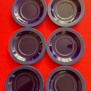 May include: A set of six circular dark blue saucers arranged in two columns of three. Each saucer features a smooth, glossy finish with a series of concentric, raised circular ridges along the outer rim, creating a uniform and simple design.