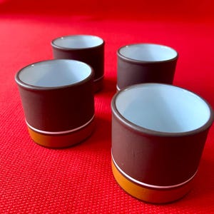 May include: Four small, brown ceramic cups with white interiors are arranged on a red surface. Each cup has a white stripe and a gold band near the base. The cups are cylindrical with a slightly flared rim.
