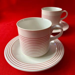 May include: Two white ceramic teacups and saucers with red horizontal stripes. The cups have a handle and sit on matching saucers. The background is a solid red colour.