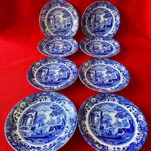 May include: A collection of eight blue and white porcelain plates. The plates showcase a scenic landscape design with trees, buildings, and water. The set includes four round plates, two oval plates, and two smaller oval plates.