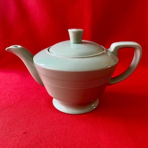 May include: A light sage green ceramic teapot featuring a smooth, glossy finish, a matching lid with a central knob, a curved spout, and a sturdy handle. The body of the teapot is decorated with three horizontal embossed rings around the centre.