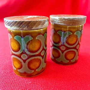 May include: Two ceramic jars with wooden lids. The jars feature a brown, orange, and green pattern. The lids are light brown. The jars are on a red surface. The jars are likely for storing spices or other small items.