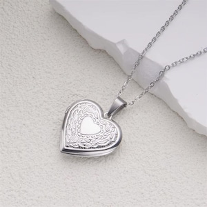 May include: A silver heart-shaped locket necklace. The locket has an engraved floral design around a smaller heart shape. The necklace chain is silver and made of small, linked pieces. The locket is displayed on a white surface.
