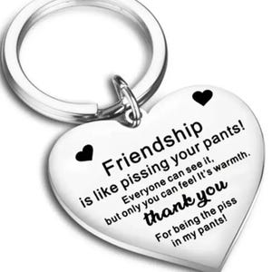 May include: A heart-shaped keyring with a silver ring. The white heart has black heart accents and text that reads "Friendship is like pissing your pants!" and "thank you".