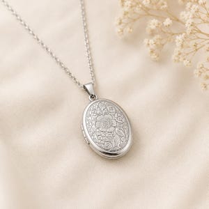 May include: A silver-toned oval locket pendant hangs from a matching delicate link chain. The front surface of the locket features an intricate, engraved floral pattern with detailed petals, leaves, and swirling vine motifs across the metal.