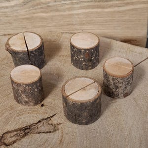 May include: Five small, rustic wooden log slices are displayed. Each has a natural bark exterior and a light-coloured, circular top. Some have a slit cut across the top, suitable for holding cards or photos. The logs are arranged on a wooden surface.