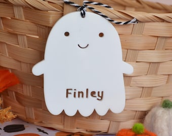 May include: A white ghost-shaped ornament featuring two small circular eyes, a curved mouth, and the name Finley engraved in brown lettering. The ornament is suspended by a black and white twisted cord tied to a light brown woven wicker basket handle.