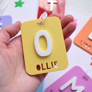 May include: A yellow luggage tag with a white letter "O" and the name "Ollie" cut out at the bottom. Other colorful tags with letters and names are visible in the background. The tag has a clear plastic loop for attachment.