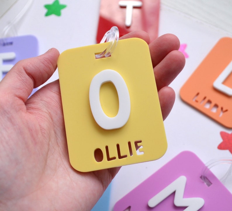 May include: A yellow luggage tag with a white letter "O" and the name "Ollie" cut out at the bottom. Other colorful tags with letters and names are visible in the background. The tag has a clear plastic loop for attachment.