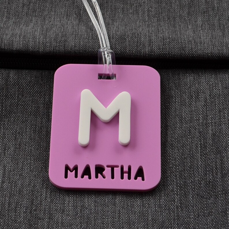 May include: A rectangular, lavender luggage tag with the white letter "M" and the name "MARTHA" cut out at the bottom. A clear plastic loop is attached at the top for securing the tag to luggage or a bag.