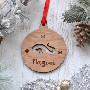 May include: A circular wooden Christmas decoration with a red ribbon. The ornament features a snake cutout and the name "Nagini" engraved beneath. Two snowflake designs are also engraved on the ornament.