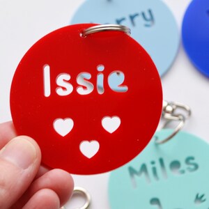 May include: A red acrylic keychain with the name "Issie" cut out, along with three heart-shaped cutouts. The keychain is round and has a silver keyring attached. Other keychains with names are visible in the background.