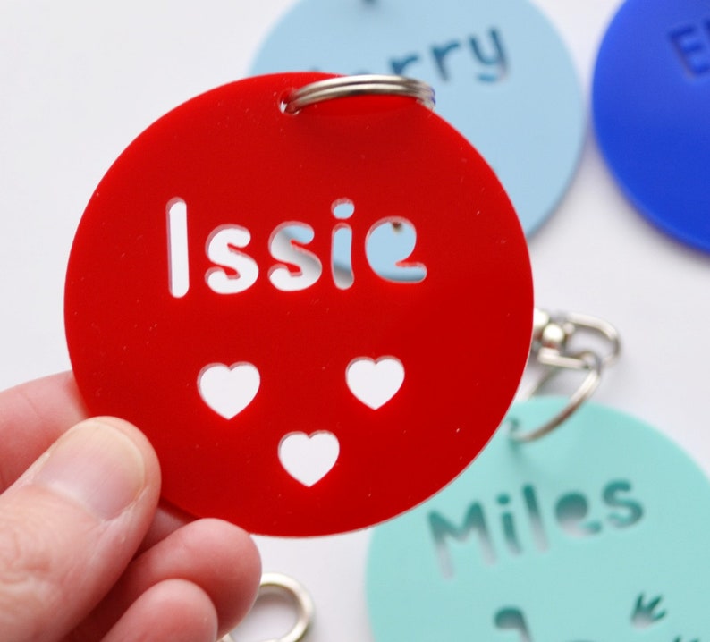 May include: A red acrylic keychain with the name "Issie" cut out, along with three heart-shaped cutouts. The keychain is round and has a silver keyring attached. Other keychains with names are visible in the background.