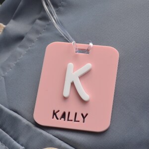 May include: A pink luggage tag with a white letter "K" and the name "KALLY" printed in white. The tag is square with rounded corners and has a clear plastic loop for attachment. The background is a gray fabric.