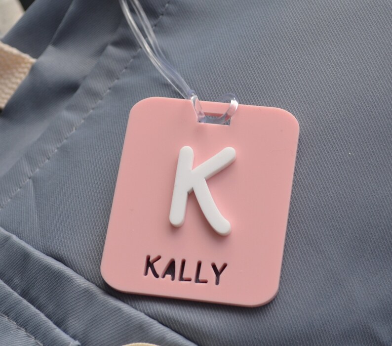 May include: A pink luggage tag with a white letter "K" and the name "KALLY" printed in white. The tag is square with rounded corners and has a clear plastic loop for attachment. The background is a gray fabric.