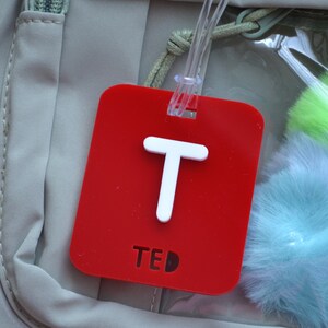 May include: A red luggage tag with a white letter "T" and the name "TED". The tag is rectangular with rounded corners and a clear plastic loop for attachment. The background includes a light-colored bag and fluffy, colorful accessories.