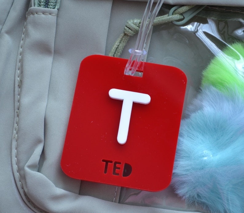 May include: A red luggage tag with a white letter "T" and the name "TED". The tag is rectangular with rounded corners and a clear plastic loop for attachment. The background includes a light-colored bag and fluffy, colorful accessories.