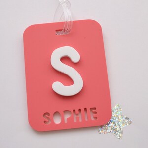 May include: A coral-colored luggage tag with a white letter "S" and the name "Sophie" cut out at the bottom. A clear plastic loop is attached at the top. A silver glitter star is next to the tag.