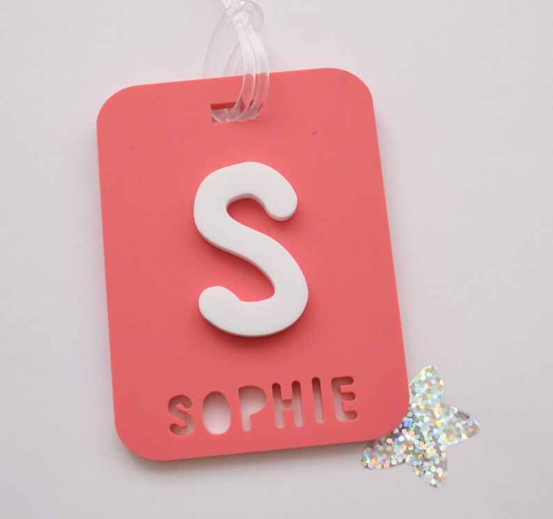 May include: A coral-colored luggage tag with a white letter "S" and the name "Sophie" cut out at the bottom. A clear plastic loop is attached at the top. A silver glitter star is next to the tag.