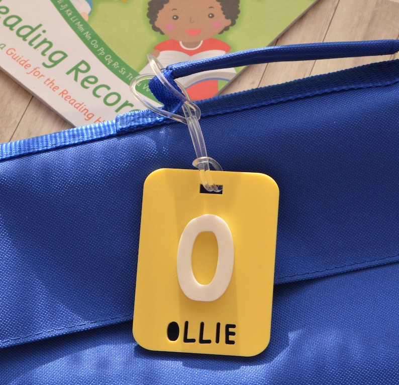 May include: A yellow rectangular tag with the letter "O" and the name "OLLIE" in black text, attached to a blue bag. The tag is hanging from a clear plastic loop. A book titled "Reading Record" is visible in the background.