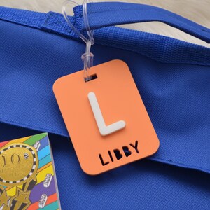May include: An orange luggage tag with a white letter "L" and the name "LIBBY" cut out at the bottom. The tag is attached to a blue tote bag with a clear plastic loop. A colorful book is visible in the lower left corner.