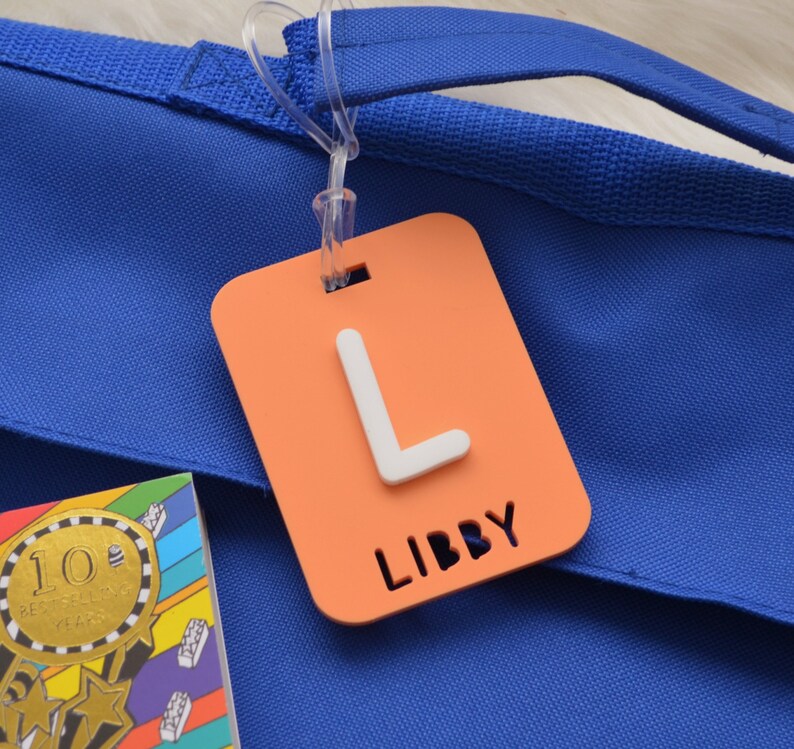May include: An orange luggage tag with a white letter "L" and the name "LIBBY" cut out at the bottom. The tag is attached to a blue tote bag with a clear plastic loop. A colorful book is visible in the lower left corner.