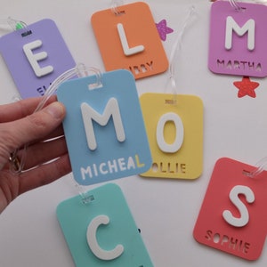 May include: A collection of colorful luggage tags in various shades of blue, yellow, orange, purple, and red. Each tag features a large white letter and a name. The tags are rectangular with rounded corners and a clear plastic loop.