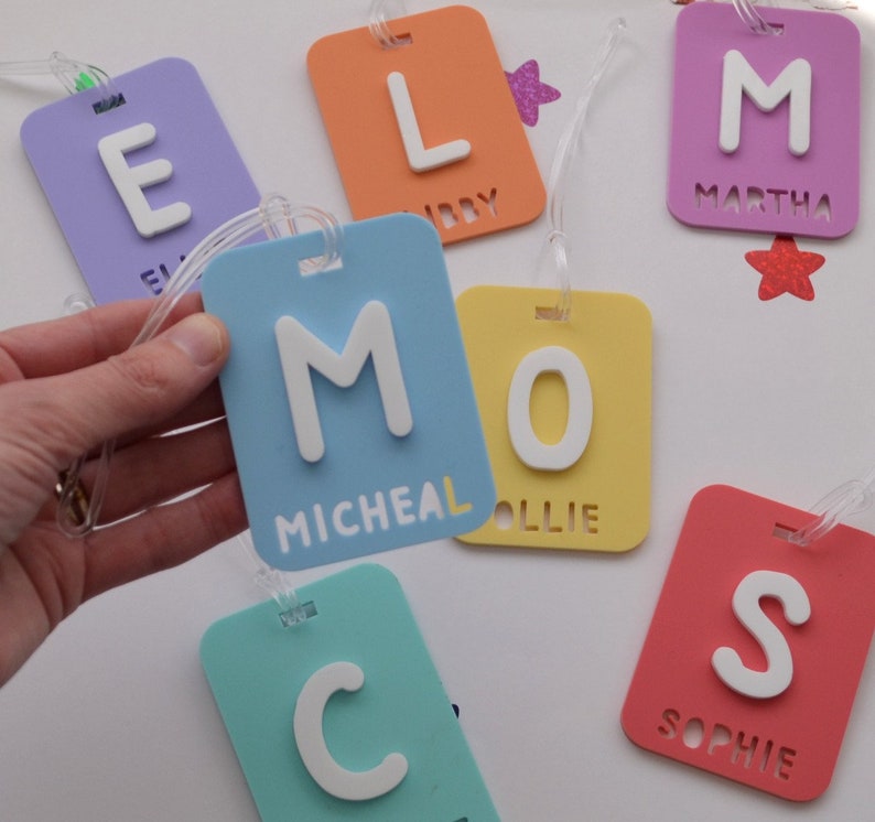May include: A collection of colorful luggage tags in various shades of blue, yellow, orange, purple, and red. Each tag features a large white letter and a name. The tags are rectangular with rounded corners and a clear plastic loop.