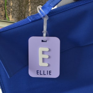 May include: A light purple luggage tag with the letter "E" and the name "ELLIE" in white. The tag is attached to a blue bag with a clear plastic loop.