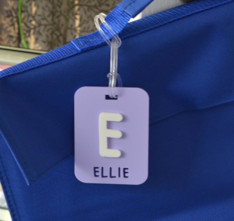 May include: A light purple luggage tag with the letter "E" and the name "ELLIE" in white. The tag is attached to a blue bag with a clear plastic loop.