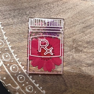 Peut inclure: Pendentif rectangulaire transparent en résine en forme de flacon de médicament. À l'intérieur, une fleur rouge vibrante est visible, ainsi que de petites fleurs séchées violettes et roses. Le contour blanc du flacon et le symbole Rx sont proéminents.