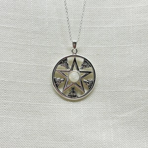 Peut inclure: Collier pendentif argenté avec un motif de pentagramme. Le pendentif présente une étoile centrale et un cadre circulaire orné de silhouettes de têtes de loup. Une petite pierre ronde blanc cassé est au centre de l'étoile.
