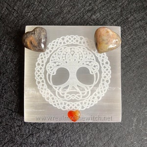 Peut inclure: Une plaque de recharge en s&eacute;l&eacute;nite blanche avec un motif d'arbre de vie et une bordure de n&oelig;ud celtique. Trois cristaux en forme de c&oelig;ur sont plac&eacute;s sur la plaque : un cristal noir et brun, un cristal rose et brun et un cristal de cornaline rouge. La plaque porte le texte "www.realmofthewitch.net" imprim&eacute; au bas.