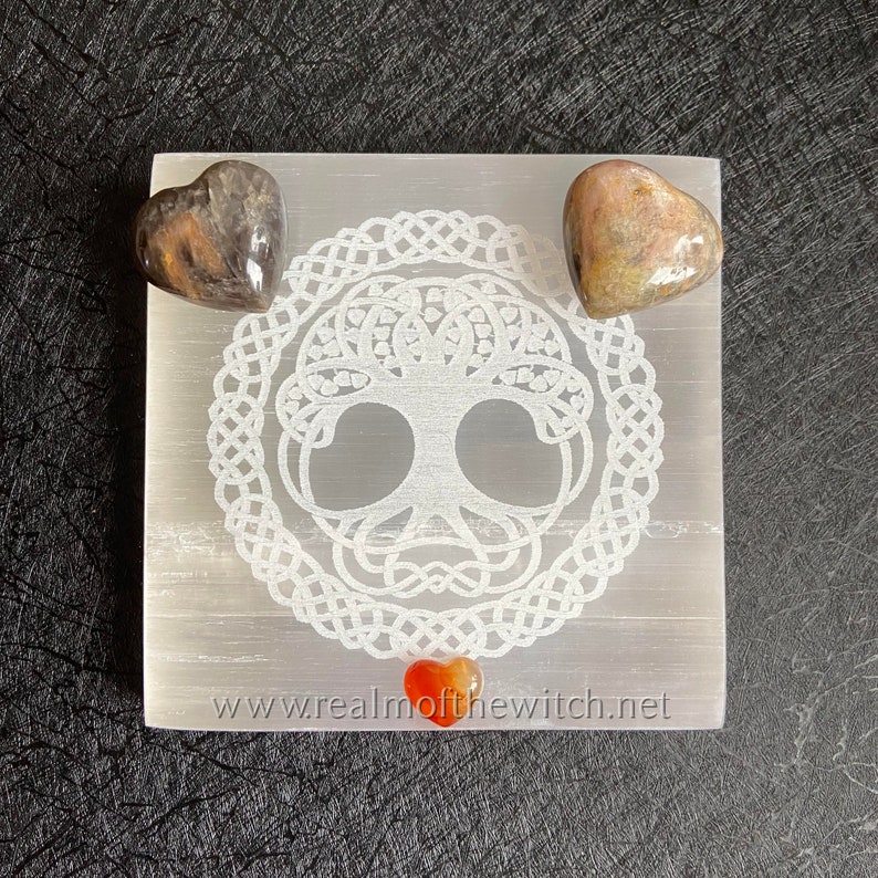 Peut inclure: Une plaque de recharge en s&eacute;l&eacute;nite blanche avec un motif d'arbre de vie et une bordure de n&oelig;ud celtique. Trois cristaux en forme de c&oelig;ur sont plac&eacute;s sur la plaque : un cristal noir et brun, un cristal rose et brun et un cristal de cornaline rouge. La plaque porte le texte "www.realmofthewitch.net" imprim&eacute; au bas.