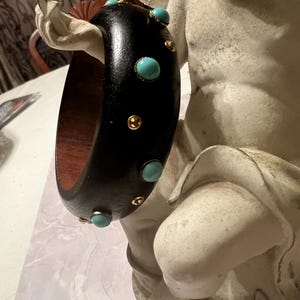 May include: A black wooden bangle bracelet with turquoise and gold accents. The bracelet has a smooth, polished finish and a contrasting reddish-brown interior. The turquoise stones are round and evenly spaced around the bracelet, with small gold accents in between.
