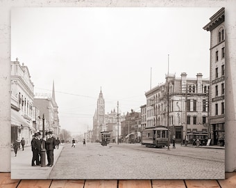 May include: A black and white photograph of a city street scene. The street is lined with buildings, and there are people walking on the sidewalk. There is a streetcar in the middle of the street.