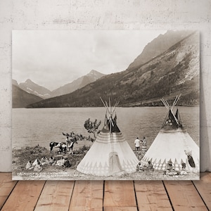 Può includere: Indiani Blackfeet accampati nel Glacier National Park, Montana. Due tipi sono installati sulla riva di un lago con montagne sullo sfondo. Le persone sono radunate intorno ai tipi.