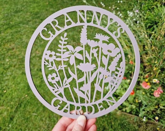May include: A round, light purple garden sign with the words "GRANNY'S GARDEN" cut out around the edge. The center features a floral design with various flower and plant silhouettes. The sign is held in front of a green background.