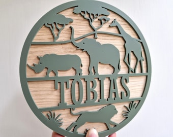 May include: A circular wooden sign with a sage green frame and a light wood centre. It features cut-out silhouettes of African animals, including a rhino, elephant, giraffe, and lion, with the name "TOBIAS" cut out in large letters.