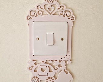 May include: A light pink decorative light switch cover featuring a cutout design of a carriage, a figure in a gown, stars, and a heart-shaped tiara top. The cover surrounds a standard white rectangular light switch with a central toggle.