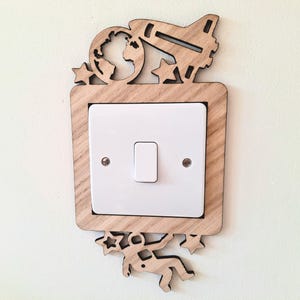 May include: Wooden light switch cover with a space theme. The cover has a rocket, stars, an astronaut, and a globe. The light switch is white and rectangular. The cover is made of light-coloured wood.
