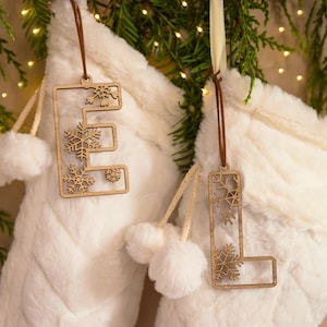 May include: Two wooden Christmas ornaments shaped like the letters "E" and "L" with snowflake cutouts. The ornaments hang from brown ribbons against a backdrop of white faux fur stockings and greenery with fairy lights.